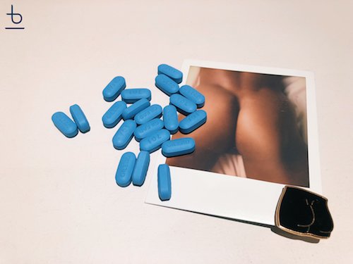 A picture of a butt posed on a bed, with a matching enamel pin and blue pills scattered across the photograph.